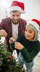 Happy young couple in Santa hats decorating a Christmas tree together. Vertical photo of a Caucasian man and Asian  woman putting up festive lights for the holiday season. Winter tradition