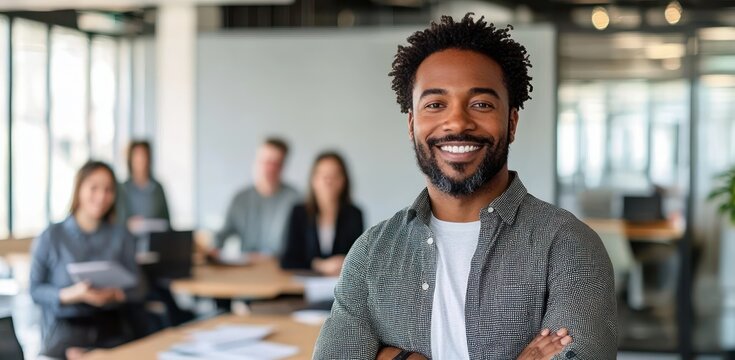 Confident smiling man standing with arms crossed in a modern office with colleagues sitting at a table behind him - Powered by Adobe