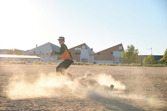 Two young baseball players making a play, one sliding and one fielding, creating a dust cloud