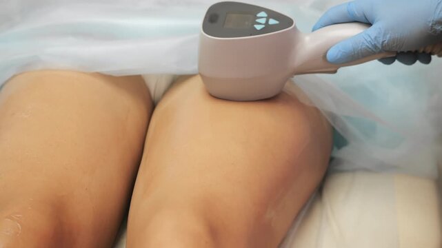 Professional muscle stimulation treatment on woman legs performed by cosmetologist in modern beauty clinic, using medical device and elastic straps indoors.