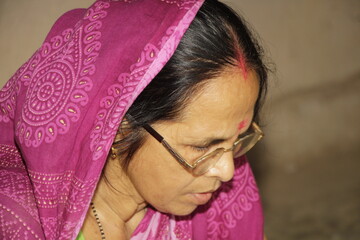 Elderly Indian woman wearing a bright pink saree with traditional patterns. She has glasses on and a red bindi on her forehead and the background is plain and neutral.