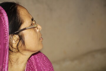 Elderly Indian woman wearing a bright pink saree with traditional patterns. She has glasses on and a red bindi on her forehead and the background is plain and neutral.