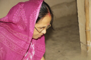 Elderly Indian woman wearing a bright pink saree with traditional patterns. She has glasses on and a red bindi on her forehead and the background is plain and neutral.