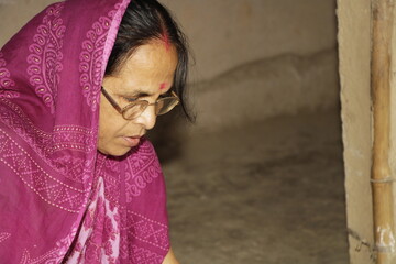 Elderly Indian woman wearing a bright pink saree with traditional patterns. She has glasses on and a red bindi on her forehead and the background is plain and neutral.