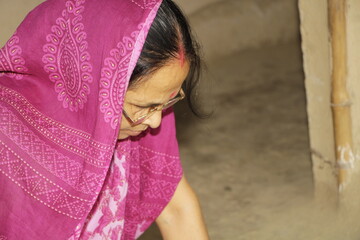 Elderly Indian woman wearing a bright pink saree with traditional patterns. She has glasses on and a red bindi on her forehead and the background is plain and neutral.