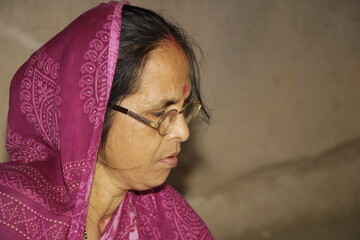 Elderly Indian woman wearing a bright pink saree with traditional patterns. She has glasses on and a red bindi on her forehead and the background is plain and neutral.