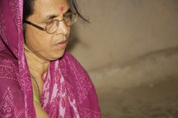 Elderly Indian woman wearing a bright pink saree with traditional patterns. She has glasses on and a red bindi on her forehead and the background is plain and neutral.