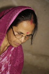 Elderly Indian woman wearing a bright pink saree with traditional patterns. She has glasses on and a red bindi on her forehead and the background is plain and neutral.
