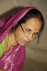 Elderly Indian woman wearing a bright pink saree with traditional patterns. She has glasses on and a red bindi on her forehead and the background is plain and neutral.