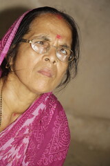 Elderly Indian woman wearing a bright pink saree with traditional patterns. She has glasses on and a red bindi on her forehead and the background is plain and neutral.