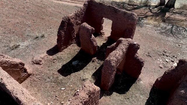 An elevated view of the ruins of an ancient or abandoned structure made of reddish mud bricks, situated on a dry bank overlooking a lake with bare winter trees