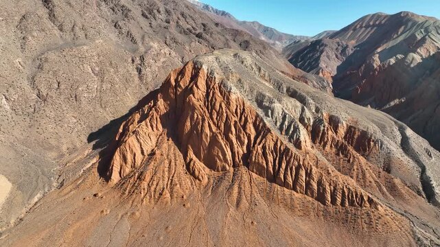 An aerial view focusing on a uniquely eroded, reddish-orange conical rock formation rising from a dry, arid plain against dark, barren mountain slopes