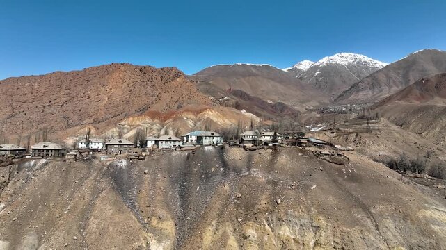 High angle view of the remote village showing rows of Soviet era residential buildings and a newer mosque, set against dramatic red-brown slopes and distant snow capped mountains