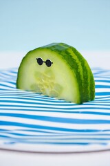 A humorous half slice of cucumber with googly eyes and eyebrows resting on a striped blue and white surface