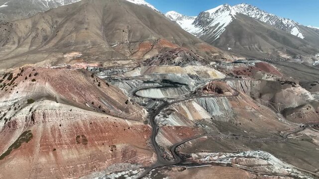 Aerial view of a massive terraced open pit or quarry in a colorful mountain landscape, featuring steep switchback roads and exposed multi-hued rock strata