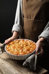 Woman with baking dish of delicious homemade apple pie at wooden table against black background, closeup