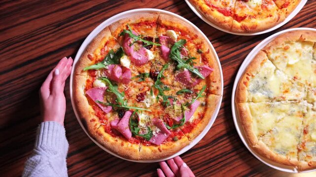 Hands place prosciutto pizza on wood, gourmet pie topped with arugula and herbs beside cheese pizza, warm closeup, crisp crust and vibrant greens, casual pizzeria mood