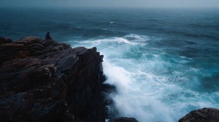 A solitary figure sits perched on a rugged cliff edge gazing out at the powerful churning waves of a stormy ocean