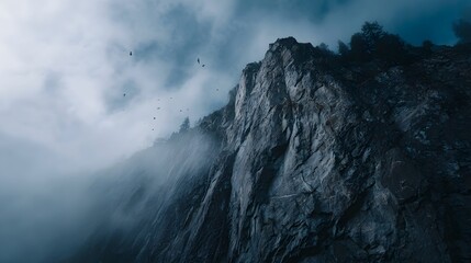 Dramatic sheer cliff face shrouded in fog with birds soaring in the cloudy sky