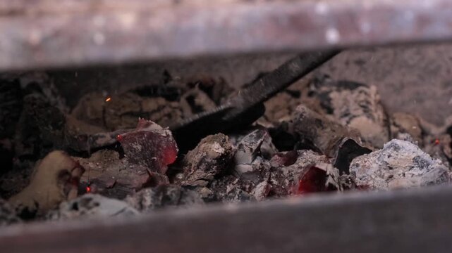 Smoldering ash and glowing embers under grill, close view of shovel turning coal, scattered burnt paper and cinder, muted light, rustic metal grate and smoky atmosphere
