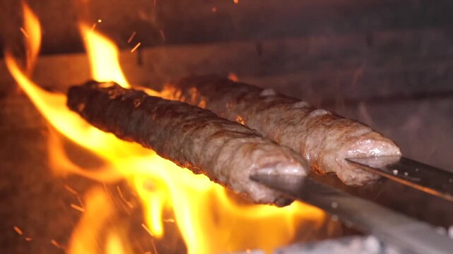 Two kebab skewers grilling over roaring flame with sparks and rising smoke, closeup of charred surface and sizzling juices in high-heat outdoor or restaurant barbecue