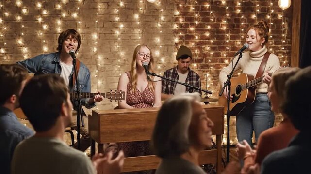 A band of musicians plays acoustic guitars and sings on stage for an appreciative audience enjoying the live performance under warm string lights.