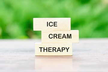 The arrangement shows three wooden blocks with ICE, CREAM, and THERAPY printed on them. It sits outdoors, on a table, with a blurred green background, creating a calming message
