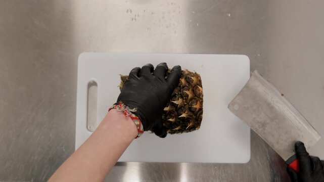 Hands slicing pineapple, Overhead perspective of hands preparing pineapple with serrated blade, Topdown view showing gloved hands halving juicy pineapple on clean white