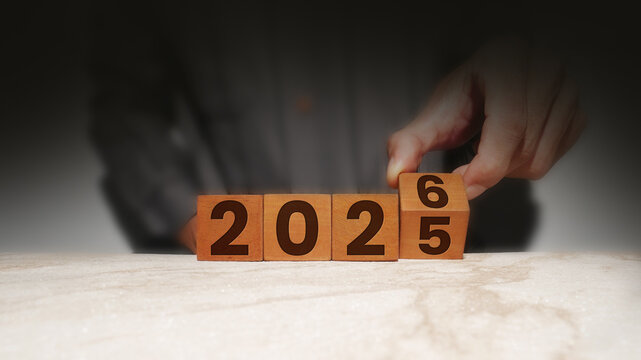 A businessman's hand in a dark, shadowy office is changing wooden blocks from the year 2025 to 2026 on a marble desk.