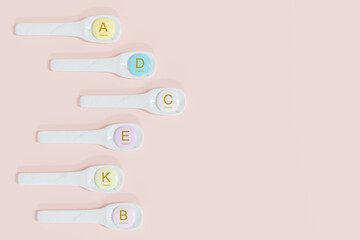 A flat lay showing six spoons with pastel-colored vitamin supplements (A, D, C, E, K, B) arranged vertically on a soft pink background. healthy supplements concept. 3d render.