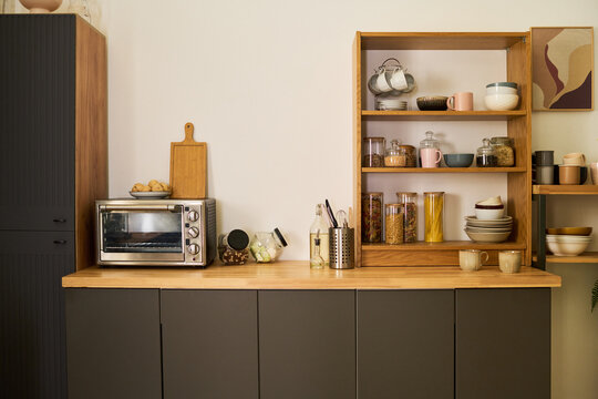 Modern kitchen countertop featuring electric toaster oven, wooden cutting board, glass jars with grains and pasta, ceramic bowls, mugs, utensils, open shelving, framed wall art