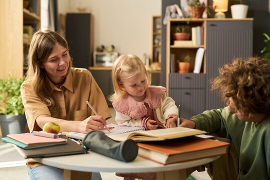 Caucasian young adult woman sitting at table helping Caucasian toddler girl with drawing while Caucasian teenager boy reading book nearby, all engaged in creative learning activity indoors
