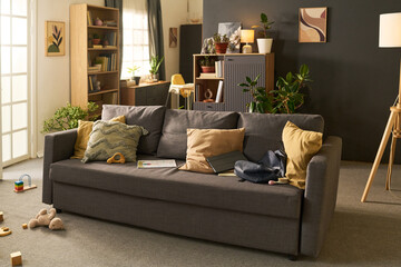 Modern living room featuring gray sofa with scattered toys, open laptop, backpack, and books...