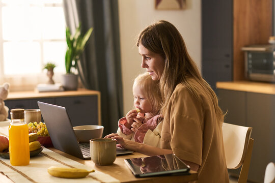 Caucasian young adult woman working on laptop while holding toddler girl on lap in kitchen, both looking at screen, breakfast items and tablet on table, natural morning light streaming in