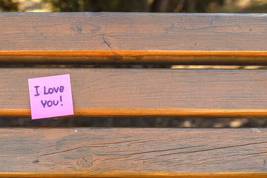 “I Love You” Note on Bench. Pink sticky note with handwritten “I love you” message on a wooden bench. Symbol of affection, care, and love communication.