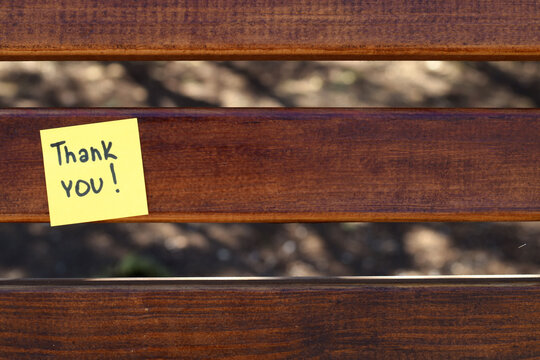 Thank You Note on Wooden Bench. A yellow sticky note with a handwritten “Thank You!” message and a smiley face attached to a wooden bench in sunlight. Perfect for expressing gratitude, positivity