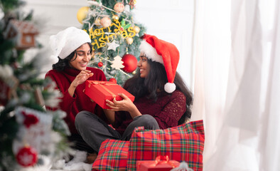 Two cheerful Indian girls exchanging Christmas gifts and smiling beside a decorated Christmas tree