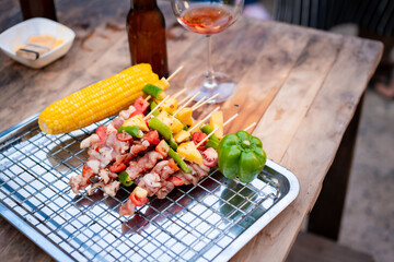 Close-up of BBQ skewers, corn, and bell pepper on a wooden table beside wine and beer. The warm string lights and blurred background create a cozy outdoor party atmosphere