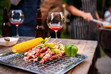 Close-up of BBQ skewers, corn, and bell pepper on a wooden table beside wine and beer. The warm string lights and blurred background create a cozy outdoor party atmosphere