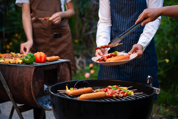 An Asian woman with friends grilling BBQ skewers, corn, and sausages outdoors. They toast with wine amid glowing string lights, creating a joyful and warm evening barbecue atmosphere