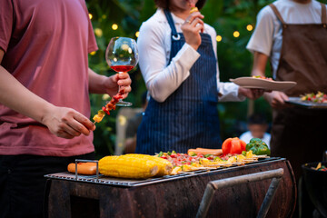 An Asian woman with friends grilling BBQ skewers, corn, and sausages outdoors. They toast with wine amid glowing string lights, creating a joyful and warm evening barbecue atmosphere