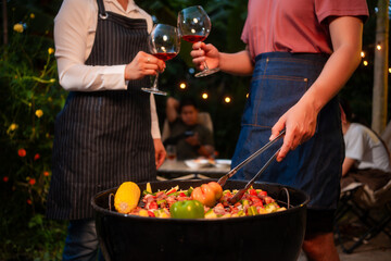 An Asian woman with friends grilling BBQ skewers, corn, and sausages outdoors. They toast with wine amid glowing string lights, creating a joyful and warm evening barbecue atmosphere
