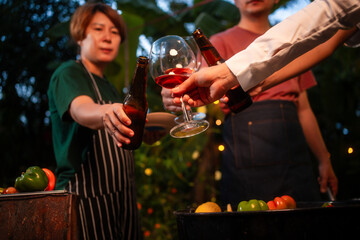 An Asian woman with friends grilling BBQ skewers, corn, and sausages outdoors. They toast with wine amid glowing string lights, creating a joyful and warm evening barbecue atmosphere