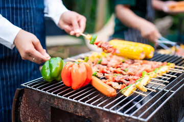 An Asian woman grills assorted barbecue skewers with meat, bell peppers, pineapple,corn outdoors. The backyard setting captures a cheerful summer cookout filled with color, warmth,delicious aromas