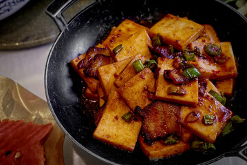 A close-up of square pieces of pan-fried golden tofu and slices of pork belly tossed in a dark, spicy chili sauce and green onions, served in a black cast-iron skillet. 