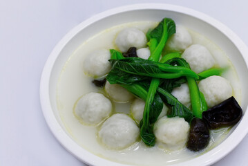 A top-down close-up of a white bowl filled with creamy fish ball soup. The dish contains smooth white fish balls, vibrant green Chinese greens (choi sum), and dark wood ear mushrooms