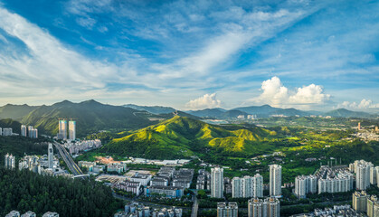 Aerial panorama of Shenzhen city edge where urban developments and highways meet lush green mountains under a dramatic cloudy sky.
