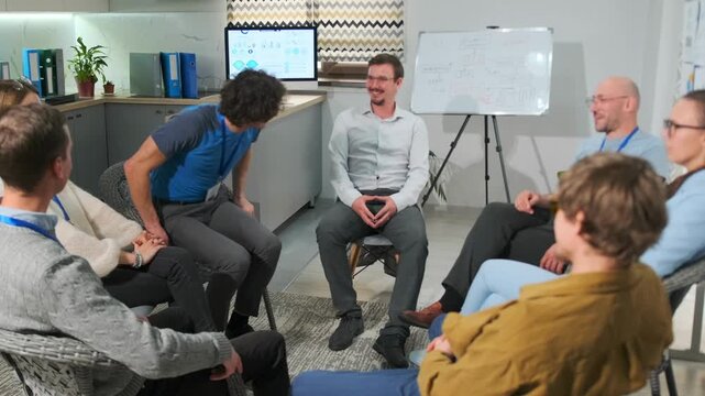 Young man in a blue T-shirt stands up at a psychotherapy meeting, showing his badge and introducing himself as part of a corporate training on trust and relationships. Colleagues applaud in support.