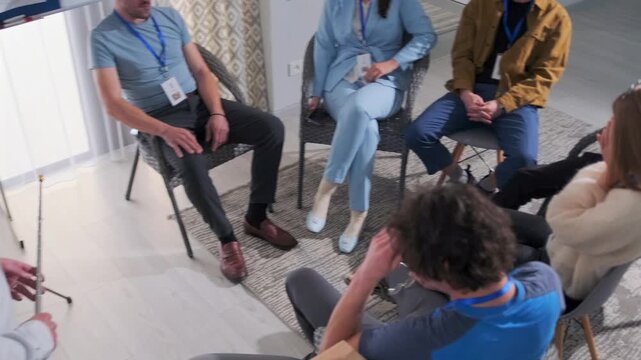 Business team sits in a circle during a coaching session. The trainer with a pointer stands by a flipchart, while employees laugh, support each other, and engage in positive team-building atmosphere.