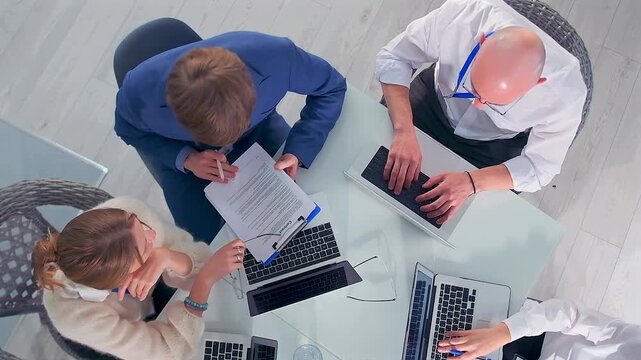 overhead client pitch around glass table, advisor presenting funding proposal to entrepreneur and senior consultant, team reviewing charts and metrics on laptop, hopeful collaborative energy in modern